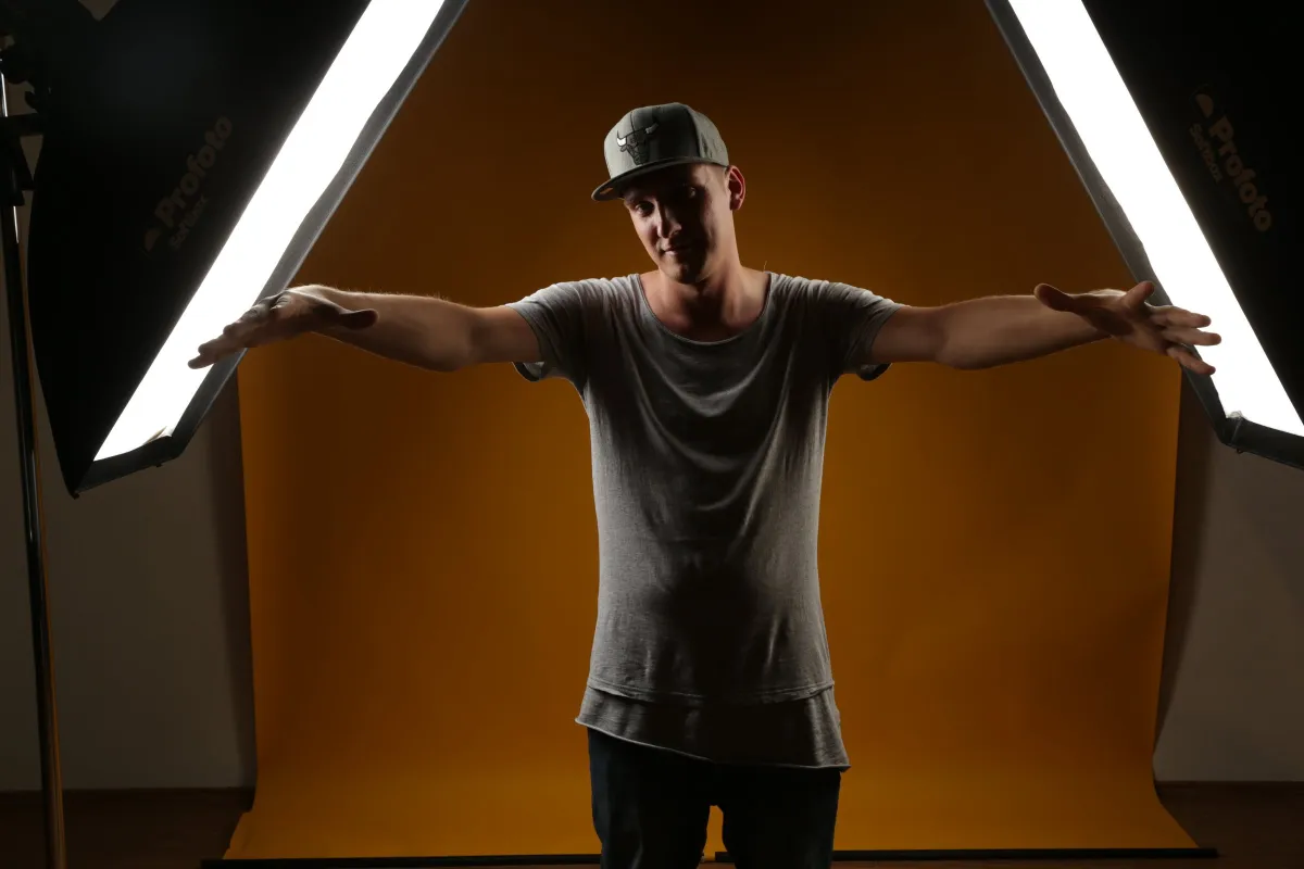DJ EMC mit Cap breitet vor orangem Hintergrund die Arme aus, links und rechts leuchten Softboxen