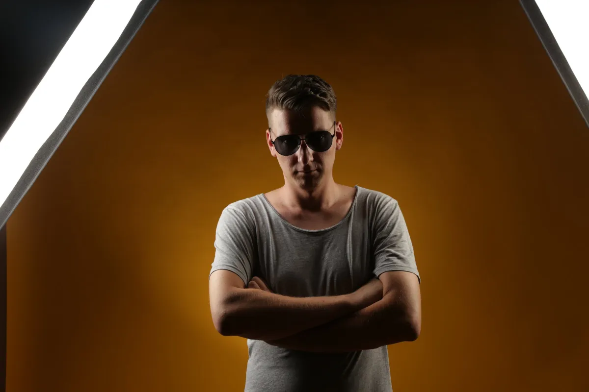 DJ EMC mit Sonnenbrille und verschränkten Armen zwischen zwei Softboxen, oranger Hintergrund