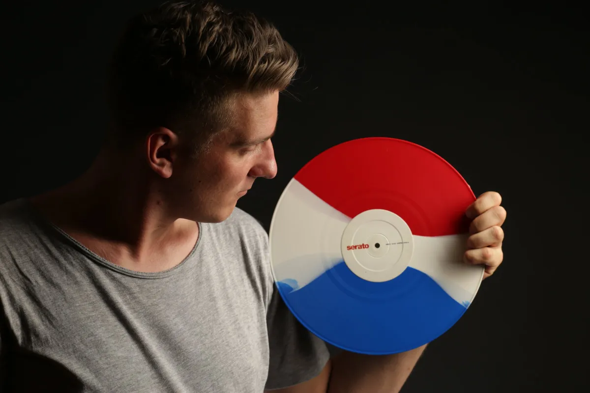DJ EMC hält eine blau-weiß-rote Serato-Vinyl neben sein Gesicht, dunkler Hintergrund