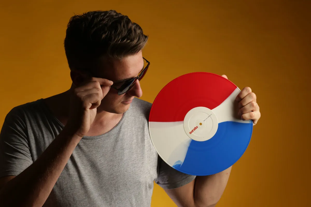 DJ EMC rückt seine Sonnenbrille zurecht und hält eine blau-weiß-rote Serato-Vinyl, oranger Studiohintergrund