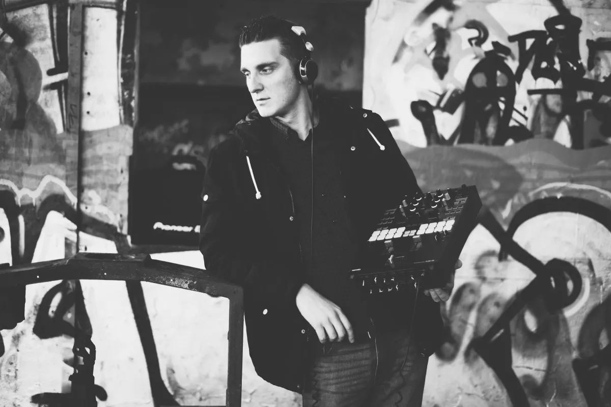 Schwarz-Weiß-Foto: DJ EMC mit Kopfhörern und DJ-Controller unter dem Arm lehnt an einem Geländer vor Graffiti
