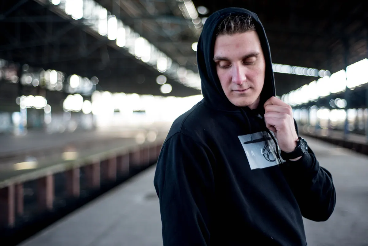DJ EMC im schwarzen Hoodie mit Kapuze auf einem Bahnsteig, Blick nach unten