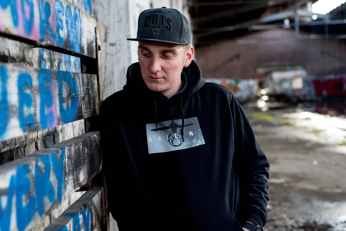 DJ EMC mit Cap und schwarzem Hoodie lehnt an einer Wand mit blauem Graffiti