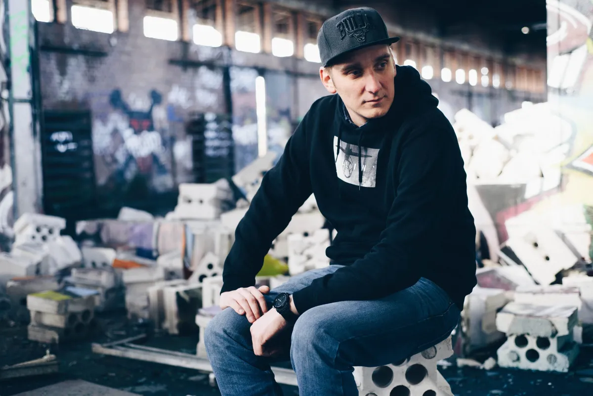 DJ EMC mit Cap und Hoodie sitzt auf Mauersteinen in einer Industriehalle mit Graffiti