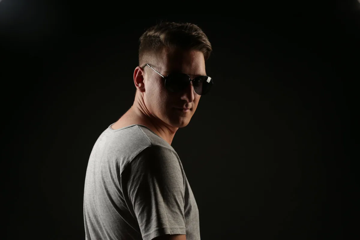 DJ EMC mit Sonnenbrille und grauem Shirt vor schwarzem Hintergrund, seitlich über die Schulter blickend