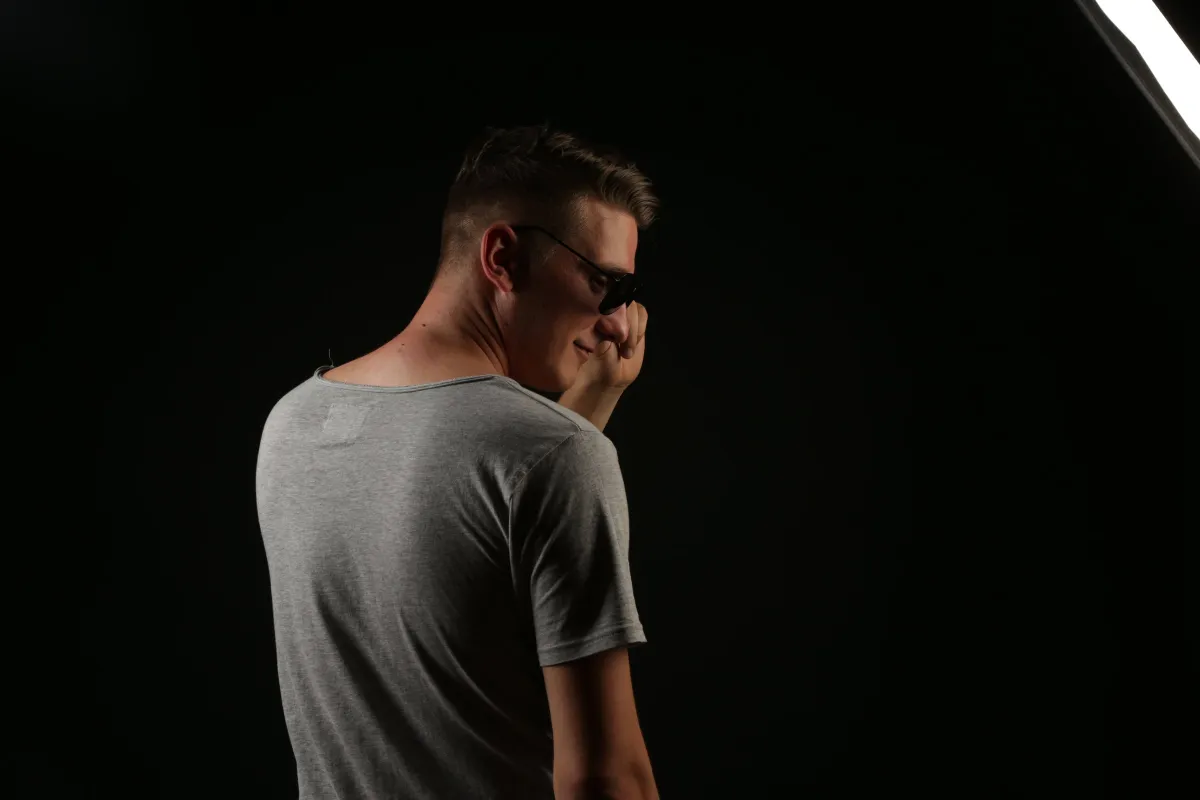 DJ EMC mit Sonnenbrille, seitlich abgewandt, die Hand am Gesicht, vor dunklem Hintergrund