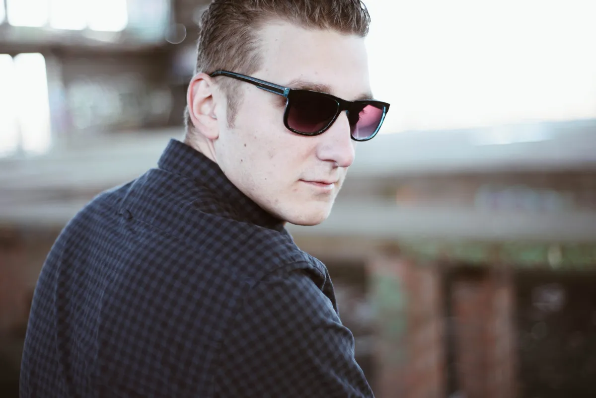 DJ EMC mit Sonnenbrille blickt über die Schulter in die Kamera, unscharfer Industriehintergrund