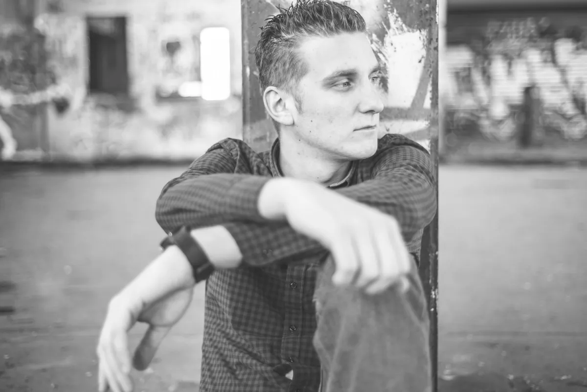 Schwarz-Weiß-Foto: DJ EMC lehnt sitzend an einem Pfeiler vor einer Industriefassade