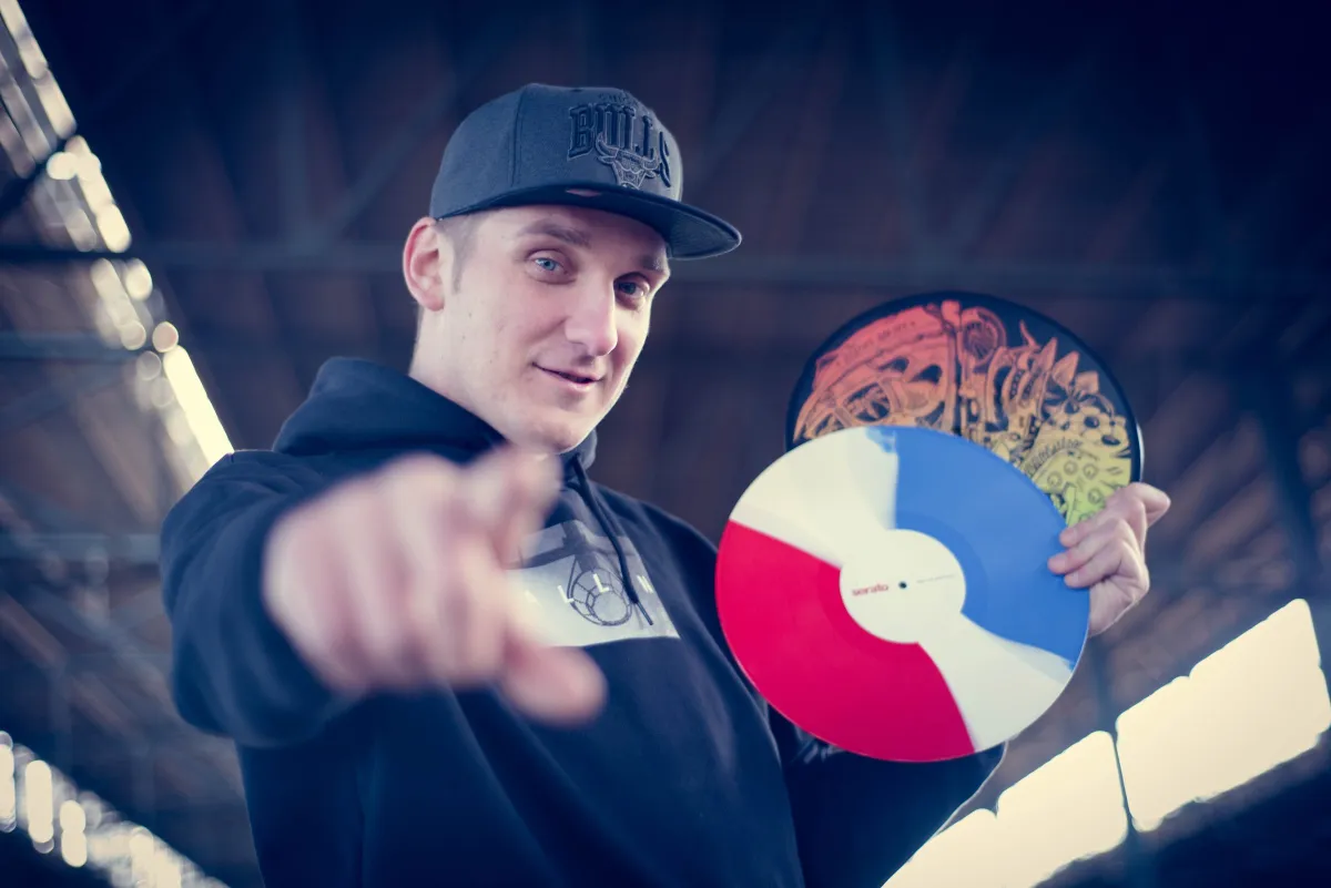 DJ EMC mit Cap und Hoodie zeigt auf den Betrachter und hält eine blau-weiß-rote Vinyl in einer Industriehalle